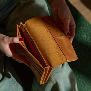 Peut inclure: Un portefeuille en cuir beige avec un rabat repliable et une fermeture à bouton-pression en métal doré. L'intérieur présente un compartiment zippé marron foncé, plusieurs fentes pour cartes et une doublure en cuir brut texturé.