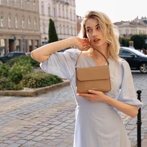 May include: A tan-colored handbag with a flap closure and a short handle. The bag is held by a person wearing a light blue dress. The background features a European city street with buildings and a car.