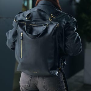 May include: A black leather backpack featuring a vertical zippered pocket on the front, gold-toned hardware, and top carry handles. The bag has adjustable shoulder straps and a minimalist design with a small gold logo printed near the bottom edge.