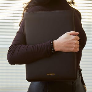 Può includere: Una custodia rettangolare per laptop in pelle nera con finitura liscia e opaca e bordi cuciti. La parola "incarne" è impressa in piccole lettere color oro vicino al centro inferiore del pannello frontale della custodia protettiva.