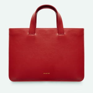 May include: A rectangular red leather handbag featuring two top handles and a smooth, minimalist exterior. The brand name "incarne" is printed in small gold lettering near the bottom centre of the bag, which has clean, stitched edges.