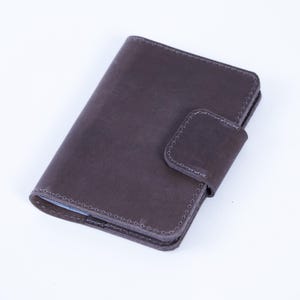 May include: A dark brown leather wallet with a rectangular shape and a flap closure. The wallet has visible stitching around the edges and a smooth, slightly textured surface. The wallet is designed to hold cards and cash.