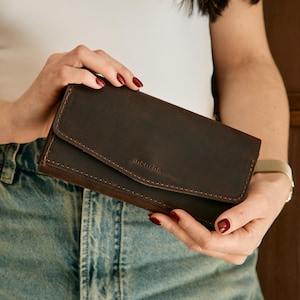 May include: A dark brown leather wallet with a rectangular shape and a fold-over flap closure. The wallet features visible stitching along the edges and the brand name Incarne embossed in the center of the front flap.