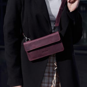 May include: A rectangular burgundy leather crossbody bag with a flap closure and a matching adjustable strap attached by silver-toned metal rings. The front of the bag features the embossed text INCARNE in a clean, sans-serif font.