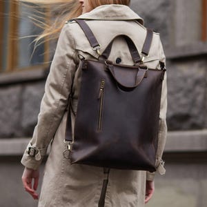 May include: A dark brown leather backpack featuring a vertical zippered pocket on the front, two top handles, and adjustable shoulder straps with brass-toned metal hardware. The bag has a rectangular shape and a smooth, matte finish.