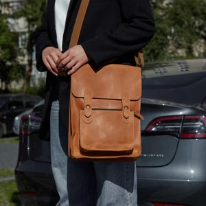 Op de afbeelding: Een bruinleren messenger bag met een klepsluiting en een voorvak. De tas heeft een schouderriem en twee gespsluitingen. De tas wordt over de schouder gedragen.