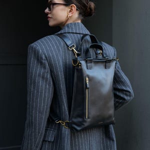 Puede incluir: Una mochila de cuero negro con un bolsillo frontal vertical con cremallera y herrajes en tono dorado, dos asas superiores y correas ajustables para los hombros sujetas con anillos y clips de metal dorado, sobre un blazer gris a rayas.