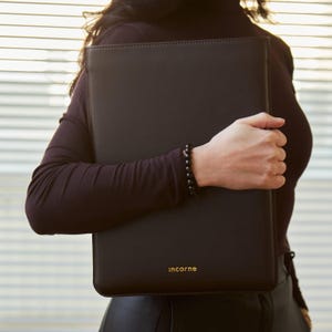 May include: A rectangular black laptop sleeve with a smooth finish and the word :incarne printed in small gold lettering at the bottom center. The sleeve features clean, stitched edges and a structured, flat design for carrying a portable computer.