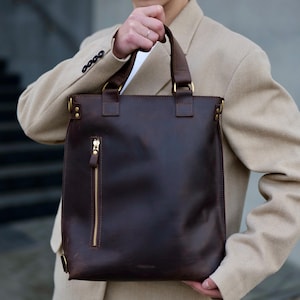 Könnte beinhalten: Eine dunkelbraune Ledertasche mit zwei Tragegriffen, einer vertikalen Reißverschlusstasche auf der Vorderseite mit goldfarbenen Beschlägen und messingfarbenen Metallringen, die die Riemen mit dem Korpus verbinden.