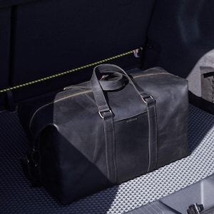Puede incluir: Una bolsa de lona de cuero negro con un asa superior y cierre de cremallera. La bolsa tiene forma rectangular y está en el maletero de un coche. La bolsa tiene una etiqueta que dice "instruktiv".