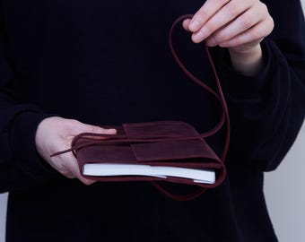 May include: A burgundy leather journal with a wrap-around closure. The journal is held open, revealing white paper pages. A long, thin leather strap secures the journal. The cover has a flap that tucks in to close the journal.