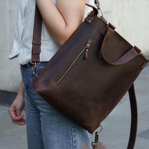 Op de afbeelding: Een donkerbruine, converteerbare leren tas met een verticaal ritsvak aan de voorzijde, twee handvatten en verstelbare schouderbanden met metalen hardware. De tas heeft een rechthoekige vorm en een gladde, matte afwerking op het leer.