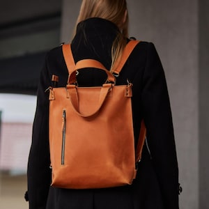May include: A tan leather backpack featuring a vertical zipped pocket on the front, two top carry handles, and adjustable shoulder straps. The bag has a rectangular shape with visible stitching details and metal hardware attachments at the top corners.
