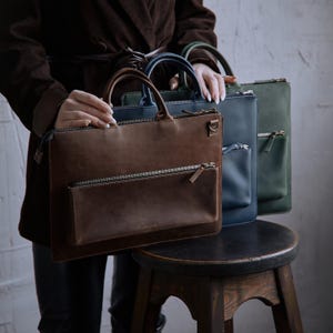 Può includere: Tre cartelle rettangolari in pelle nei colori marrone, blu navy e verde scuro. Ogni borsa è dotata di un manico superiore, uno scomparto principale con cerniera e una tasca frontale orizzontale con cerniera. La borsa marrone riporta la scritta incaro.