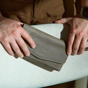 May include: A gray leather wallet with the word "incarne" embossed on the front. The wallet is rectangular with a flap closure and visible stitching. The wallet is held by a person.