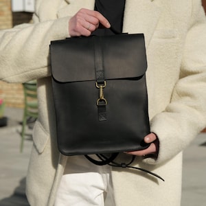 May include: A black leather backpack with a flap closure and a brass-colored clasp. The bag has a top handle and adjustable straps. The backpack is held up by a person wearing a cream-colored coat and white pants.