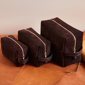 May include: Three dark brown suede travel bags of varying sizes. Each bag has a gold-colored zipper and a small leather handle. The bags are arranged on a brown leather surface.