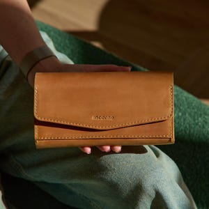May include: A tan leather wallet with a rectangular shape and a fold-over flap closure. The word "incarne" is embossed in small letters on the front flap. The wallet features visible stitching along the edges and a smooth, matte finish.