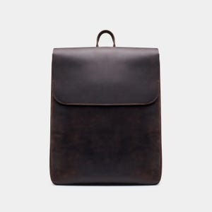 May include: A dark brown leather backpack with a flap closure and a small handle at the top. The bag has a rectangular shape with rounded corners and a smooth, textured surface. The backpack is designed for carrying items.