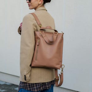May include: A tan leather backpack featuring a vertical zipped side pocket, two top handles, and adjustable shoulder straps with gold-toned metal hardware. The bag has a structured, rectangular shape and is worn over a tan blazer and plaid shirt.