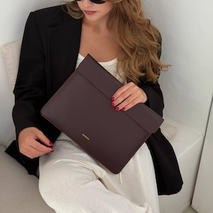May include: A dark brown rectangular laptop sleeve with a smooth finish and a flap closure. The word "incarne" is printed in small gold lettering on the lower front centre of the sleeve, which is held horizontally against a black blazer and white top.