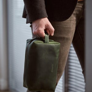 May include: A rectangular olive green leather toiletry bag featuring a top handle, a side zipper closure with a leather pull tab, and the embossed brand name "INGGINE" on the front surface. The bag has a smooth matte finish and structured shape.