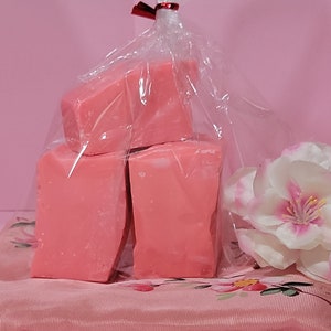 May include: Three pink bars of soap wrapped in clear plastic and tied with a red ribbon. The soap is sitting on a pink and white floral fabric.