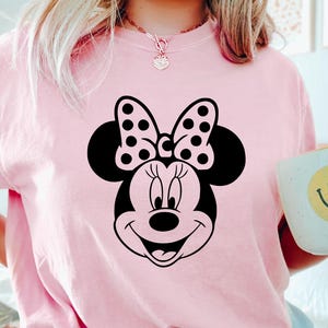 Puede incluir: Camiseta rosa claro con un gr&aacute;fico de Minnie Mouse en blanco y negro. El dise&ntilde;o incluye la cara, el lazo y las orejas de Minnie. Tambi&eacute;n se ve una taza blanca con una carita sonriente amarilla.