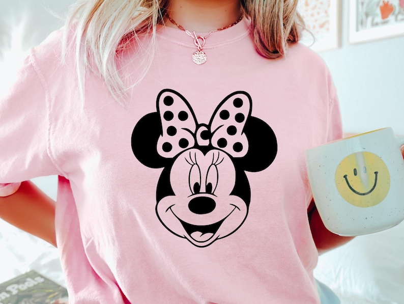 Puede incluir: Camiseta rosa claro con un gr&aacute;fico de Minnie Mouse en blanco y negro. El dise&ntilde;o incluye la cara, el lazo y las orejas de Minnie. Tambi&eacute;n se ve una taza blanca con una carita sonriente amarilla.