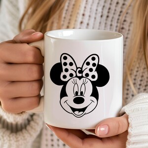 Puede incluir: Taza de cer&aacute;mica blanca con un gr&aacute;fico en blanco y negro de Minnie Mouse. El dise&ntilde;o incluye el lazo ic&oacute;nico de Minnie con lunares y una cara sonriente. La taza es sostenida por una persona.