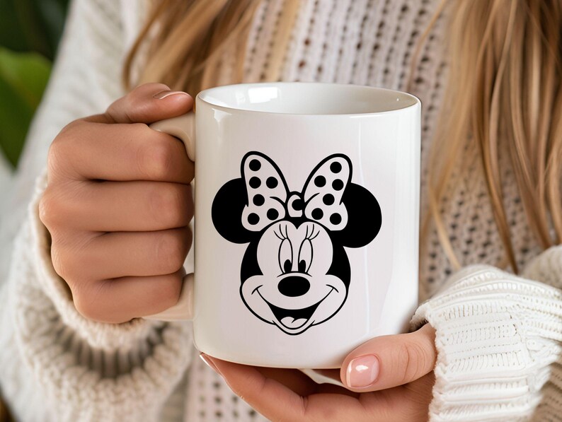Puede incluir: Taza de cer&aacute;mica blanca con un gr&aacute;fico en blanco y negro de Minnie Mouse. El dise&ntilde;o incluye el lazo ic&oacute;nico de Minnie con lunares y una cara sonriente. La taza es sostenida por una persona.