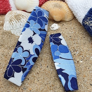 May include: Two blue and white floral patterned hair clips on a sandy beach background.