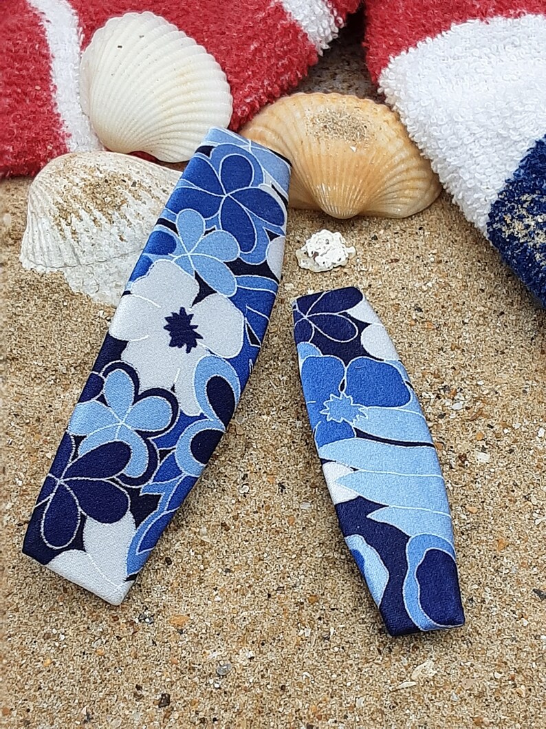 May include: Two blue and white floral patterned hair clips on a sandy beach background.