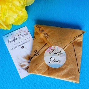 May include: A brown paper package with a white circle sticker that says "Pacific Grace" in cursive. The package is sitting on a blue surface. The sticker has a watercolor floral design.