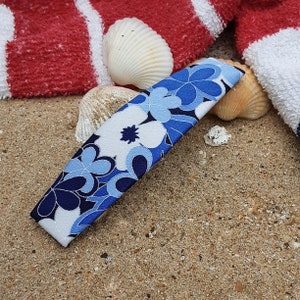 May include: A blue and white floral patterned hair clip with a curved shape. The clip is lying on a sandy surface with seashells.