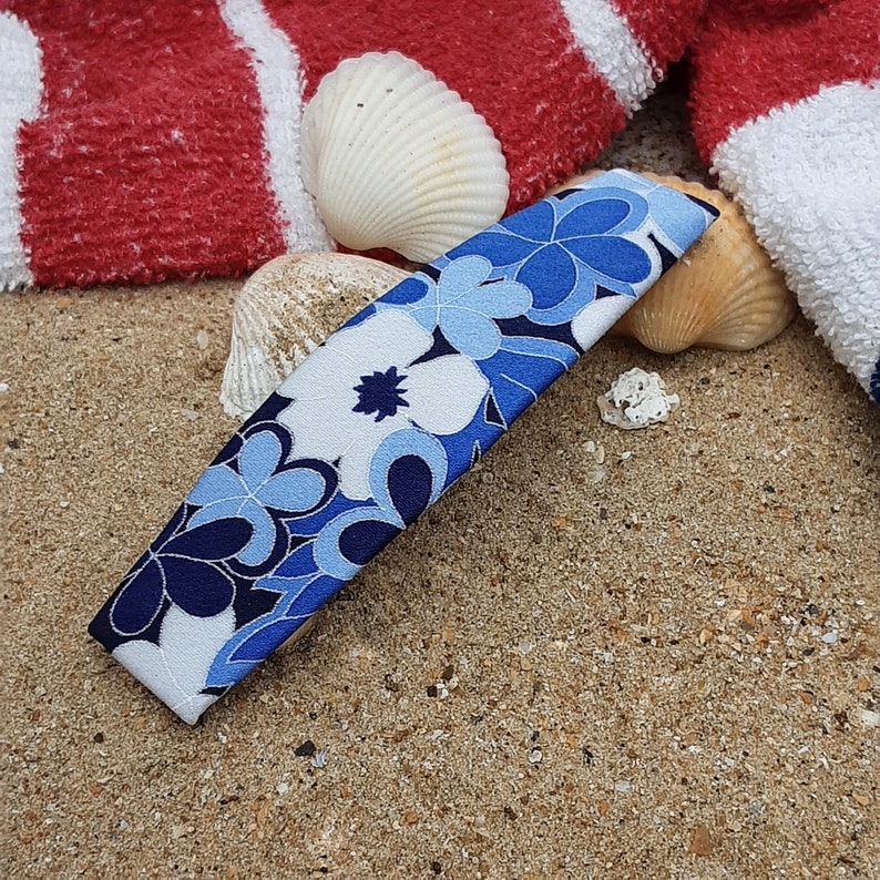 May include: A blue and white floral patterned hair clip with a curved shape. The clip is lying on a sandy surface with seashells.