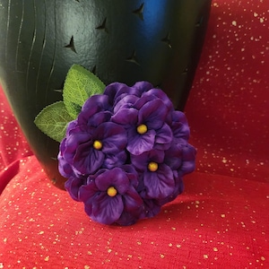 May include: A cluster of deep purple artificial flowers with yellow centers and green leaves. The flowers are set against a dark green vase with star-shaped cutouts, on a red background.