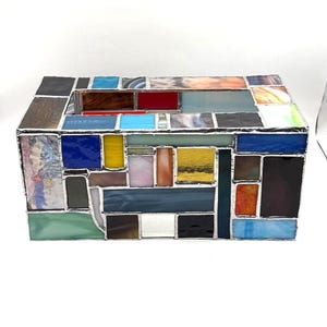 May include: A rectangular tissue box cover constructed from numerous small, multi-colored stained glass pieces joined by silver-toned metal solder. The glass panels feature a variety of textures and colors, including blue, yellow, red, and white.