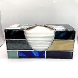 May include: A rectangular tissue box cover constructed from various pieces of stained glass joined with silver-toned metal solder. The glass panels feature a mix of colors including deep blue, light blue, beige, black, and white in a geometric pattern.