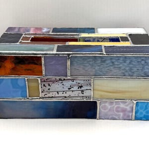 May include: A rectangular stained glass box with a variety of colors, including blue, orange, purple, and white. The glass pieces are arranged in a brick-like pattern and are held together with silver-colored metal. The box is a decorative item.