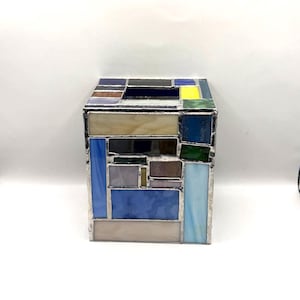 May include: A square stained glass tissue box cover constructed from various rectangular and square glass panels in shades of blue, white, grey, green, and yellow, joined together with silver-colored metal solder lines in a geometric pattern.