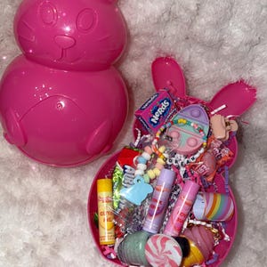 May include: A bright pink Easter bunny-shaped container filled with colorful candies, lip gloss, and a bracelet. The bunny container is next to a matching bunny-shaped basket filled with treats. The image is taken on a white, fluffy surface.