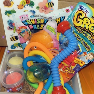 May include: A box filled with colorful sensory toys. Includes a blue and red extendable tube, a package of "Squishy Palz", stickers, and small containers of play sand and clay. The box is on an orange surface.