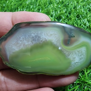 Puede incluir: Una piedra de ágata pulida, de forma ovalada, con un patrón en capas de tonos verdes, blancos y marrones. La superficie de la piedra es lisa y reflectante, mostrando bandas y remolinos naturales. El ágata se sostiene en una mano.