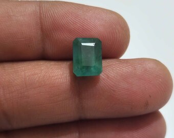 May include: A rectangular emerald gemstone with a vibrant green hue. The gem is cut with a step-cut facet design, showcasing its clarity and brilliance. The emerald is held against a neutral background, highlighting its color and shape.