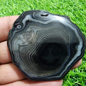 May include: A polished, circular agate stone with concentric bands of black, gray, and white. The natural stone has a smooth, glossy surface and a unique pattern, showcasing the beauty of natural formations. The stone is held in a hand.