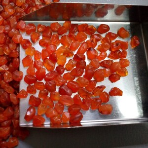 K&ouml;nnte beinhalten: Nahaufnahme eines Metalltabletts gef&uuml;llt mit kleinen, rauen, orange-roten Karneol-Edelsteinen.