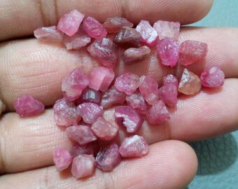 May include: A collection of small, rough-cut pink gemstones. The stones vary in shape and size, with a range of pink hues from light to deep rose. The gemstones are displayed in a hand, showcasing their natural, unpolished state. Ideal for jewelry making.