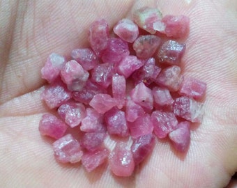 May include: A handful of rough-cut, pink tourmaline gemstones. The raw crystals vary in size and shape, displaying a range of pink hues, from light to deep rose. The gemstones are held in an open palm, showcasing their natural, unpolished state.
