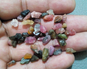 May include: A collection of small, raw gemstones in various colors, including pink, green, brown, and black. The gemstones are irregularly shaped and appear to be natural, with a rough texture. The stones are held in a hand.